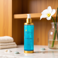 Blue skincare bottle with gold cap on a wooden surface with a white flower and towels in the background